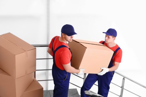 Two professional removal workers dressed in red shirts, blue overalls, and matching blue caps are inside a residential building, engaged in a house move on a stairway landing. The worker on the left is holding a large cardboard box with white gloves, while the worker on the right stabilizes the box, also wearing white gloves. In the background, several stacked cardboard boxes of varying sizes are placed against a plain white wall, indicating an organized packing process. The staircase features a black handrail with horizontal metal bars, and the scene is brightly lit with natural light, creating a clear and professional atmosphere. The setting suggests they are preparing to move household items, including boxes and possibly smaller furniture, as part of a house removals service in Twickenham or nearby areas. The image reflects the careful handling and logistical coordination typical of local house relocation efforts by Twickenham Man with Van, emphasizing the organized and professional approach to residential moves.