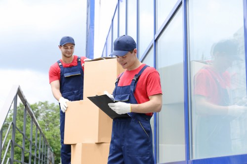 Two removal workers from Twickenham Man with Van are engaged in loading cardboard boxes onto a van parked outside a modern building with large glass windows. The worker in the foreground, wearing a navy cap, red t-shirt, blue overalls, and white gloves, is holding a clipboard and appears to be checking the packing list or inventory. Behind him, the second worker, also dressed in red and blue work attire with gloves, is carrying a stack of flattened cardboard boxes, preparing to load them into the vehicle. The scene takes place on a cloudy day, with the building's reflective glass facade to the right and a residential street or garden visible in the background to the left, suggesting a suburban or urban location in Twickenham. The environment indicates an organized effort in house removals or relocations, with focus on careful handling of household items and boxes. This image visually complements professional moving services in London boroughs, specifically in Twickenham, within the context of house removals or property transfers.