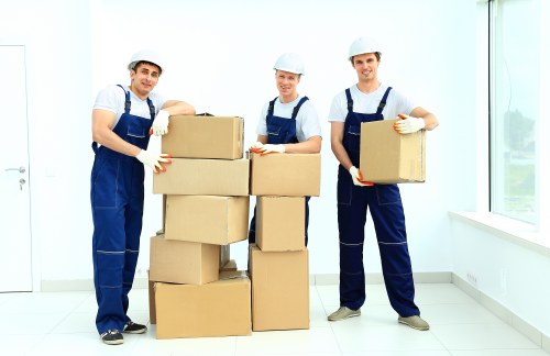 Three professional movers dressed in matching blue overalls, white shirts, and white protective helmets are inside a bright, spacious room with white walls and large windows that let in natural light. They are standing on a clean, white tiled floor, each holding a cardboard box. The boxes are uniformly brown, with some stacked in a neat formation, and appear to be filled with household items for a house removal. The man on the left is holding a box with a slight smile, the middle mover is standing behind a stack of three boxes, and the man on the right is carrying a box towards the camera, all wearing work gloves. The room has a minimalist appearance with no visible furniture or personal belongings, emphasizing the moving process. The scene subtly references residential property in Twickenham or nearby areas, aligning with professional removals and house relocation services provided by Twickenham Man with Van. The natural lighting highlights the clarity and professionalism of the team preparing for a local move within the London suburb.