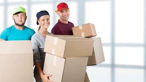 Three professional movers from Twickenham Man with Van are indoors, smiling and holding large cardboard boxes ready for a house removal in Twickenham. The background features large windows allowing natural light to illuminate the scene, suggesting a modern residential property. The first mover on the left wears a teal shirt and a bright green cap, holding a medium-sized box covered with packing tape. The middle mover, a woman, is dressed in a grey shirt and a dark blue cap, holding a large, flat package, with a friendly expression. The third mover, a man, is wearing a maroon shirt and a red cap, holding two boxes stacked one on top of the other, both sealed with packing tape and appearing sturdy. The group is positioned centrally in the image, with the boxes occupying the foreground, indicating an active packing or loading process. The scene emphasizes careful handling of household belongings, preparing for a smooth relocation in the Twickenham area, consistent with the removals services offered by Twickenham Man with Van.