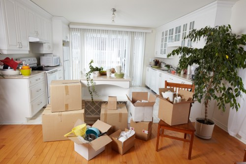 The image depicts a bright, modern kitchen with white cabinetry and wooden flooring in a residential property, subtly indicating a location within Twickenham. The kitchen area is filled with various household items and moving boxes, some open, revealing packaging materials, household goods, and small appliances such as a kettle and a microwave. Several cardboard boxes, some packed and sealed, others partially filled with items, are placed both on the floor and on a wooden dining chair, suggesting an ongoing house move or packing process. A white dining table is situated near large windowed sliding doors, which allow natural light to illuminate the space; the doors are covered with sheer white curtains. To the right, a large leafy potted plant adds greenery to the room. In the background, the kitchen countertops hold miscellaneous objects including a potted plant, and the overall scene conveys the organized chaos typical of home removals, with Twickenham Man with Van providing professional house removal services in the local area.