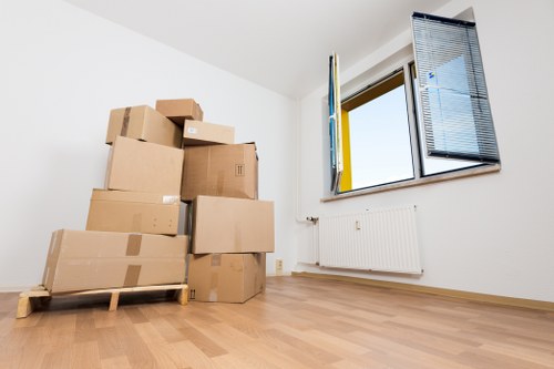 The image depicts a sparsely furnished room with white walls and a light wooden floor, likely in a residential property within Twickenham or nearby in the postal area. In the foreground, several cardboard boxes of various sizes, some sealed with brown packing tape, are stacked on a small wooden pallet, ready for removal or transportation. The boxes appear to contain household items and are neatly arranged, suggesting the process of packing or unpacking in progress. The room features a large window on the right side with partially open blue venetian blinds, allowing natural daylight to illuminate the space. Below the window, a white radiator is fixed to the wall. The environment is clean and minimally decorated, with no furniture or personal belongings visible other than the packing boxes, aligning with a house or flat prepared for a move. This scene illustrates typical contents and conditions seen during house or flat relocations in the Twickenham area, serviced by Twickenham Man with Van, a specialist in local removals and transport within the house moving industry.