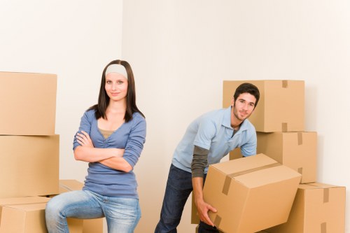 A young woman and a young man with light skin are in a room with plain white walls, surrounded by several large cardboard moving boxes of various sizes. The woman, seated on a box, has long dark hair and is dressed in a blue long-sleeved top and light blue jeans, with her arms crossed and a neutral expression. The man, standing and slightly bending forward, has short dark hair and is wearing a light blue shirt with darker sleeves, jeans, and is holding a large cardboard box, appearing to be engaged in a house removal or relocation process. The boxes are stacked on the floor and one is positioned behind the woman, with some boxes on the right side arranged in a neat cluster. The environment is well-lit with soft indoor lighting, suggesting an interior space prepared for moving, consistent with domestic house removals in Twickenham or surrounding areas.