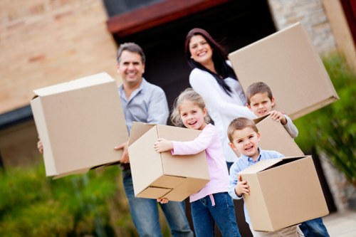 A family of four, including two adults and two children, standing outdoors in front of a residential house during daylight, holding several cardboard boxes of different sizes and shapes, with some boxes partially open. The father, on the left, is wearing a light blue shirt and jeans, smiling while holding a large box. The mother, on the right, has long dark hair, wearing a white top and dark trousers, smiling as she holds a medium-sized box. The young girl in the middle is wearing a pink long-sleeved top and jeans, hugging a smaller box to her chest, smiling directly at the camera. The young boy, standing in front, is wearing a blue shirt and beige trousers, holding a box with a cheerful expression. The background shows a brick house with an open garage door, with greenery and a driveway visible, indicating a suburban setting. The scene reflects a typical house move in Twickenham, engaging Twickenham Man with Van’s removals services, emphasizing the household items and family involvement typical of local removals in the area.