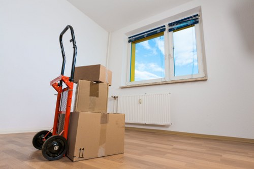 The image shows a minimally furnished room in a residential property, likely in the Twickenham area, with three large cardboard moving boxes stacked near a white wall and a black and orange hand truck positioned beside them. The boxes are closed, with some tape visible on the flaps, and vary in size, with the largest at the bottom and smaller boxes stacked above. The room features light wood laminate flooring and white painted walls, with a white radiator mounted beneath a double-glazed window. The window has a white frame and partially opened blue and yellow blinds, revealing a bright sky with some clouds outside. Natural daylight illuminates the space, casting soft shadows on the floor. The scene appears to be during a house move, associated with professional removals services in Twickenham, as indicated by the context of the webpage for pricing and quotes at twickenhammanwithvan.co.uk. The overall environment suggests an early stage of a house relocation process, with minimal furniture or personal belongings in view, emphasizing packing and logistics preparations for a house move.