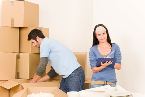 A man and a woman preparing boxes for a house move, with the man on the left bending over a partially opened cardboard box, handling packing materials or items, while the woman on the right stands holding a clipboard or tablet device, looking at the camera with a neutral expression. The background features a plain white wall with several stacked cardboard boxes of various sizes, some sealed with packing tape and others open. The scene takes place in a residential interior, possibly a hallway or living room, with a light, neutral atmosphere suitable for house removals services in Twickenham or nearby areas. The image highlights typical household packing activity involving cardboard boxes, packing supplies, and the preparation for a move. The overall setting reflects a professional yet casual environment, consistent with house removals handling personal belongings.