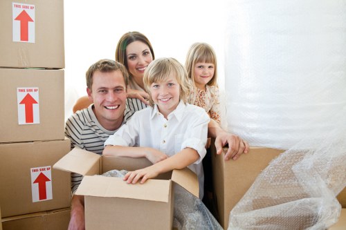 A family of four, including two children and two adults, inside a house surrounded by moving boxes, some of which are large cardboard containers with red directional arrows and branded with 'This Side Up.' The family members are smiling and appear to be in the process of packing or unpacking, with the children sitting on a cardboard box in the foreground. The children are holding or leaning on the boxes; the boy has light brown hair and is wearing a white shirt, while the girl has blonde hair and is dressed in patterned clothing. The woman, with long dark hair, and the man, with short light brown hair, are positioned behind the children, both smiling affectionately. The background shows a white wall with soft natural lighting, typical of a residential interior, and the scene suggests a move or relocation within Twickenham or nearby areas in southwest London, captured during daytime in a typical home setting. The environment emphasizes the household items involved in a house removal process, with an atmosphere of organization and anticipation, consistent with a professional removals service such as Twickenham Man with Van.