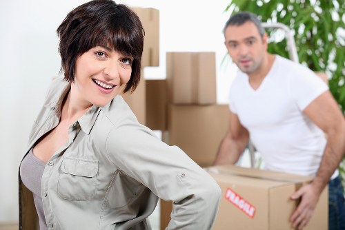 A woman with short dark hair and a light-colored shirt smiling at the camera while standing in a residential interior space, with a man in a white t-shirt in the background carrying a cardboard box, part of a house removal process. Behind them are several stacked cardboard boxes of varying sizes, some with labels, placed on the floor and near a staircase. The room features neutral walls, and natural light filters in through nearby windows, creating a bright environment suitable for house removals in Twickenham or surrounding areas. The scene captures a professional and organized atmosphere, indicative of a local house moving service handling household furniture, cartons, and personal belongings, with furniture and packing materials visible as part of the moving process.