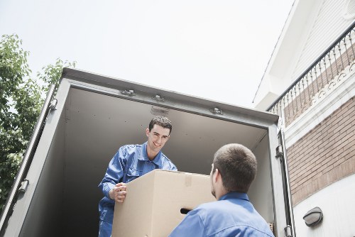 Two movers from Twickenham Man with Van, dressed in blue uniforms, unloading a large cardboard box from a removals van parked outside a residential property in Twickenham. The worker inside the van is smiling and handing the box to another mover standing outside, whose back is partly visible. The interior of the van shows a clean, metallic floor and sides, with some additional boxes visible behind the main package. The scene is set on a residential street with the upper floors of a house visible, featuring a brick wall, white cladding, and decorative balcony railing. Natural daylight illuminates the scene, highlighting the texture of the cardboard box, the fabric of the uniforms, and the exterior house details. The image captures the process of a professional house removal, emphasizing careful handling and efficient service typical of Twickenham-based removals specialists operating within the local area, including the TW postcode district.