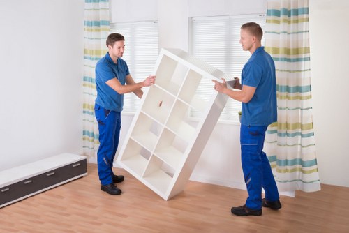 Two male movers dressed in blue uniforms are carefully lifting and repositioning a white wooden bookshelf with multiple compartments inside a bright, well-lit room. The bookshelf has a smooth finish and is being carried between the two workers, who are positioned on either side to ensure stability. The room features light wooden flooring and white walls, with large windows allowing natural daylight to fill the space. Behind the men, there are striped curtains with shades of blue, green, and beige, framing the windows. On the left side of the image, a sleek black heating unit is positioned near the floor, and the background shows a residential interior typical of homes in Twickenham or the surrounding areas. The scene clearly depicts house removals professionals managing furniture in a clean, organized environment, reflecting the careful handling of household items during a move or redeployment, consistent with services offered by Twickenham Man with Van in the area of house removals and sustainability efforts.