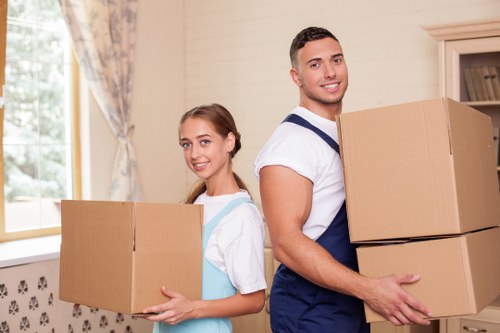 A young man and a young woman, both smiling, are standing inside a residential property in Twickenham, holding and carrying large cardboard moving boxes. The man, dressed in a white t-shirt and blue overalls, is positioned in the foreground on the right, holding two boxes, one stacked on top of the other. The woman, dressed in a light blue top, stands slightly behind him on the left, also holding a single larger cardboard box. Behind them, a cream-colored wall with floral curtains pinned at the sides and a window with a view of trees can be seen, indicating natural daylight illuminating the scene. The environment suggests a house interior, possibly a living room or hallway, within a typical UK home. The image illustrates professional house removals, with the presence of boxes and furniture being prepared for moving, relating to services offered by Twickenham Man with Van in the Twickenham postcode area, as part of their removals service category.