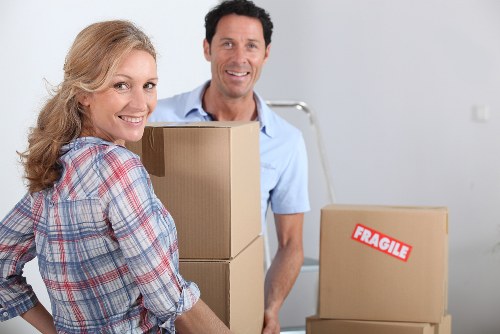 A smiling woman with curly blonde hair and a plaid shirt stands in the foreground holding brown cardboard boxes, one of which is labeled 'FRAGILE,' while a man with dark hair and a light blue polo shirt stands behind her, also smiling, in a room with light-colored walls. In the background, there are additional boxes and a metallic hand truck, indicating a house removal or moving process. The scene appears well-lit with natural or ambient lighting, suggesting a residential interior, possibly in Twickenham or nearby in the UK. The image captures the preparation stages of a professional removals service by Twickenham Man with Van, with furniture and belongings being packed for relocation within a house, emphasizing the careful handling of household items during the move.