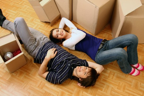 Two young women with dark hair are lying on a wooden floor amidst cardboard moving boxes in what appears to be a residential interior, possibly a living room or hallway in Twickenham. One woman, wearing a white top and blue jeans, is resting with her head on her hands and eyes closed, while the other, dressed in a dark striped shirt and light grey trousers, is lying on her back with her head turned slightly to the side and eyes open. The cardboard boxes vary in size, some open and containing household items such as a red and silver bauble and a cloth, indicating a recent or ongoing move. The background shows a partially visible stairway and a white wall, suggesting the interior of a residential property. The scene captures a moment of rest during house moves, with the arrangement of boxes, casual posture, and home setting subtly reflecting house removals and relocation services in the Twickenham area, associated with Twickenham Man with Van.