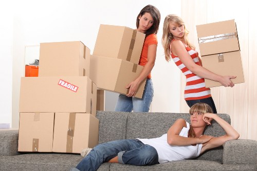 The image shows two young women involved in a home relocation process within a residential interior, standing behind a gray sofa that is surrounded by several brown cardboard moving boxes of various sizes, some sealed with packing tape and one marked with a 'Fragile' label. One woman, in a red and white striped dress, is holding a smaller box on her hip, while the other, in an orange top and blue jeans, is carrying a larger box with both hands. A young boy is lying on the sofa in the foreground, wearing a white t-shirt and blue jeans, resting his head on his hand with a thoughtful or bored expression. The background features a light-colored wall with a closed door, and faint natural daylight filtering into the space, indicating a typical living room setting in a suburban house, possibly in Twickenham or nearby in southwest London, as part of household removals or moving services provided by Twickenham Man with Van.
