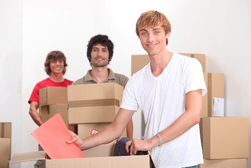 A young man with light brown hair, wearing a white t-shirt, is smiling and holding a pink envelope while standing next to a cardboard box on a table, surrounded by other packed cardboard boxes in a bright, indoor environment. Behind him, two other individuals, a woman in a red top and a man with dark hair and a beard in a beige shirt, are also smiling and holding cardboard boxes, indicating a house removal or relocation setting. The background shows a plain white wall with some placed boxes, suggesting an ongoing packing process typical of residential moves in the Twickenham or nearby London area. The scene captures the organized activity of a professional removals team, with visible cardboard boxes of various sizes, some sealed with packing tape, and the participants prepared for carrying household items. The professional setup and focus on packing and moving materials reflect company services specializing in residential relocations within the local community, supporting a smooth house move for clients in the Twickenham postcode area.