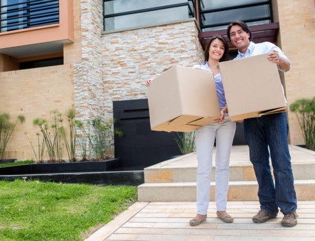 In the foreground of the image, two individuals stand outside a modern residential building in Twickenham, each holding two large cardboard boxes, likely for house relocation. The woman, positioned slightly to the left, has shoulder-length dark hair and is dressed in light grey trousers and a purple blouse. The man, to the right, has short dark hair and is wearing a white shirt with dark trousers. They are smiling and facing the camera, with the boxes partially obscuring their bodies. Behind them, the house features a contemporary design with a brick and stone facade, displaying a mix of beige, brown, and grey tones, and a large dark window or door on the upper level. To the left, a well-kept lawn with small plants and a landscaped border adds greenery, while a tiled pathway leads to the entrance stairs. The overall scene suggests a professional house moving or removals service, such as Twickenham Man with Van, in a residential area near postcode TW1 or TW2, with the environment illuminated by natural daylight, emphasizing a clean and organized outdoor setting typical of local house removals in Twickenham.