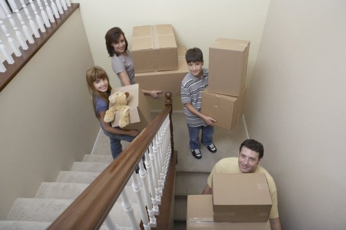 A family of four preparing to move within a residential property, seen on a staircase landing area. The two young girls, one with long red hair and the other with curly brown hair, are holding cardboard boxes and a teddy bear, standing near the top of the staircase. The boy with short dark hair is carrying a medium-sized box, while an adult man with short dark hair and a beard is seated on the staircase steps, also holding a box. The staircase has a wooden handrail with white balusters, and beige carpeted stairs lead down to a lower level. Behind the family, there are several stacked cardboard boxes of various sizes, some taped and ready for removal. The walls are painted a light neutral colour, and a window with wooden blinds can be seen on the left side, allowing natural light into the space. The scene depicts an organized moving process typical of house removals, with household belongings prepared for transport, reflecting a suburban setting in Twickenham or nearby areas in southwest London.