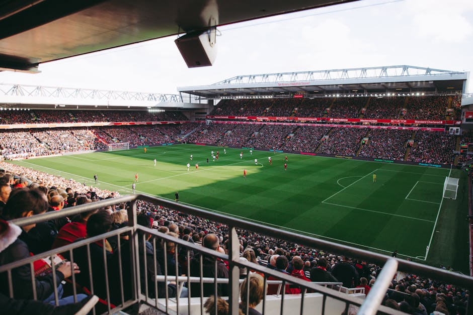 A view of a packed football stadium from a seated position near the stands, showing a well-maintained grass pitch with players actively engaged in a match. The field is surrounded by rows of spectator