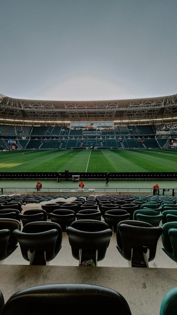 A view of a packed football stadium from a seated position near the stands, showing a well-maintained grass pitch with players actively engaged in a match. The field is surrounded by rows of spectator